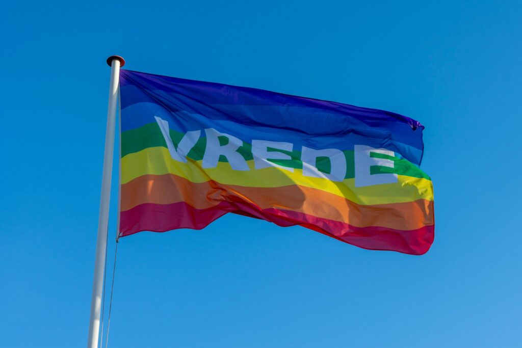 A colorful rainbow flag with 'VREDE' waving against a clear blue sky, symbolizing peace and unity.
